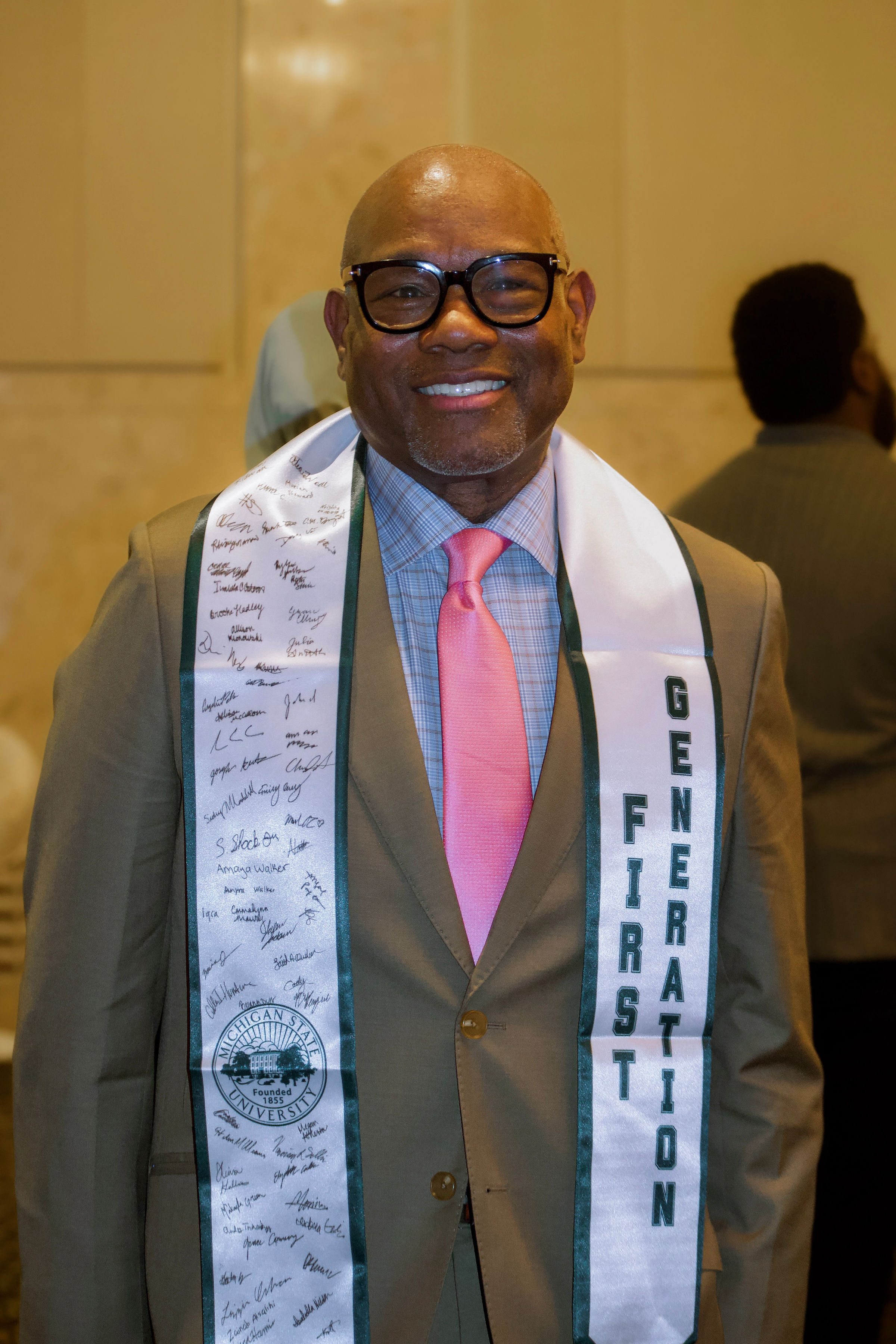 Mike Alston, wearing glasses and dressed in a tan jacket over a blue plaid button-up shirt and pink tie, smiles while sporting his own white First-Gen graduation stole. Alston’s stole has the signatures of first-gen students completely covering the side of the sash hanging down from his right shoulder.