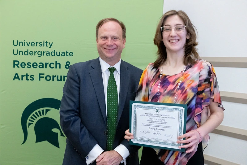 Two individuals stand next to one another smiling and facing toward the camera; they are standing in front of a green backdrop for MSU's UURAF. The person on the right is holding a large white certificate to the camera.
