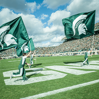 Students running across Spartan stadium field holding large spartan flags